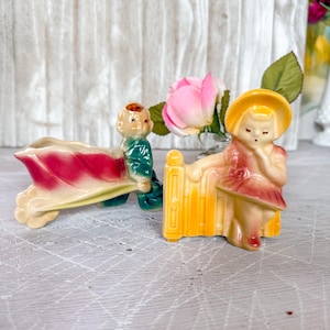 Puede incluir: Dos figuras de cerámica vintage. Una representa a un niño empujando una carretilla con un diseño rojo y crema. La otra muestra a una niña apoyada en una valla amarilla, con un vestido rojo y un sombrero amarillo. Una rosa rosa está en el fondo.