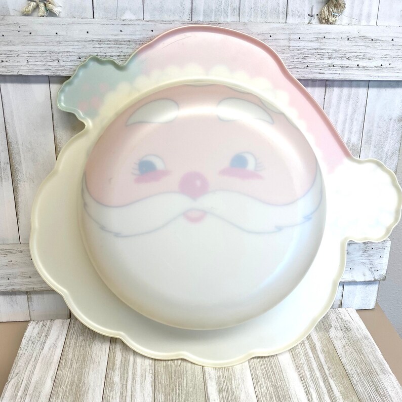 May include: A white plastic serving platter shaped like Santa Claus's face. The platter has a pink face with blue eyes and a white beard.