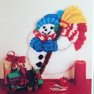 Könnte beinhalten: Ein weißer Schneemann mit einem blauen Hut und einem rot-weißen Schal, der einen gelben Besen hält. Der Schneemann hat schwarze Augen und einen braunen Mund. Der Schneemann ist von Weihnachtsdekoration umgeben, darunter eine rote Kerze, eine grüne Geschenkbox mit einer roten Schleife und ein roter Apfel.