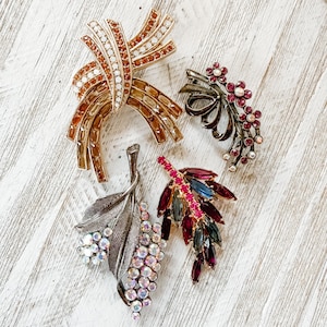 Könnte beinhalten: Eine Sammlung von Vintage-Broschen. Eine Brosche hat ein bandartiges Design mit klaren und bernsteinfarbenen Kristallen. Eine andere ist ein Blumenspray mit roten Kristallen. Eine dritte ist ein Blatt mit irisierenden Kristallen. Alle sind aus Metall.