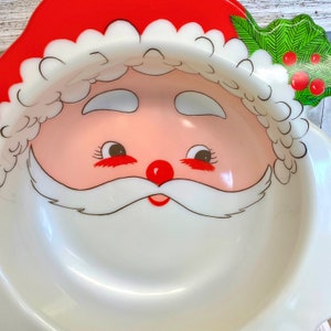 May include: White ceramic bowl shaped like Santa Claus's face. The bowl has a red hat, white beard, and black eyes. The bowl is perfect for serving holiday treats.
