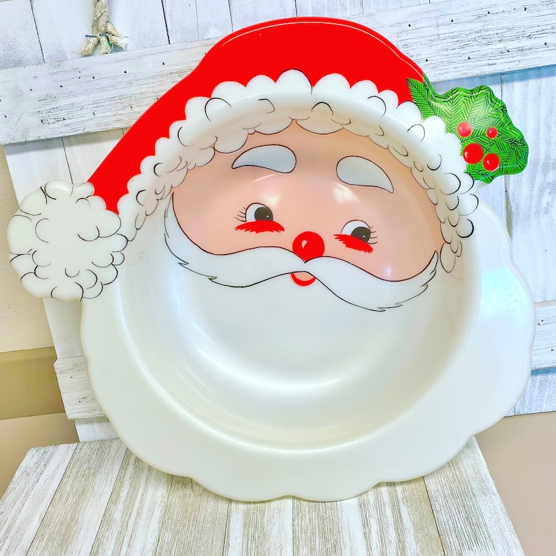 May include: A white plastic Santa Claus serving bowl with a red hat, white beard, and black eyes. The bowl has a green holly sprig with red berries on the side.