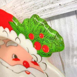May include: A white ceramic Santa Claus dish with a red hat and green holly sprig with red berries. The dish is perfect for serving snacks or treats during the holiday season.