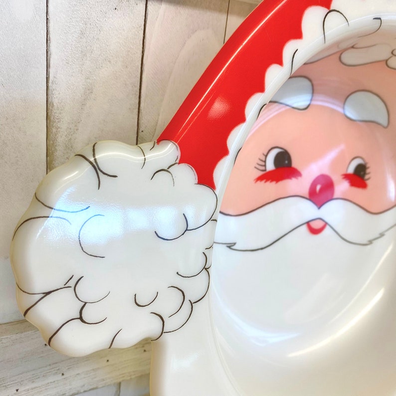 May include: A white ceramic Santa Claus serving dish with a red hat and white fur trim. The dish is shaped like Santa's face and has a white beard and black eyes.