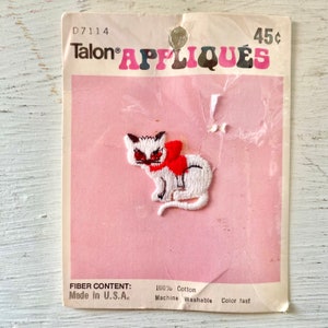 May include: White embroidered iron-on applique of a cat wearing a red bow. The applique is on a pink card with the text "Talon APPLIQUÉS" and "45c". The card also includes the text "Fiber Content: 100% Cotton, Made in U.S.A., Machine Washable, Colour fast."