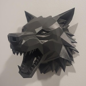 Op de afbeelding: Zwarte, geometrische wolfskop wanddecoratie met een open mond en zichtbare tanden. Het kunstwerk is bevestigd aan een witte muur en toont een modern, hoekig ontwerp. De vacht van de wolf is getextureerd.
