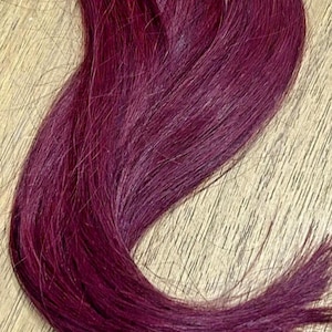 Puede incluir: Un mechón de extensiones de cabello largo y liso en un color rojo burdeos profundo y vibrante. El cabello es suave y brillante, con un tono uniforme de color vino oscuro en todas las hebras, con una pequeña etiqueta blanca visible en la parte superior.