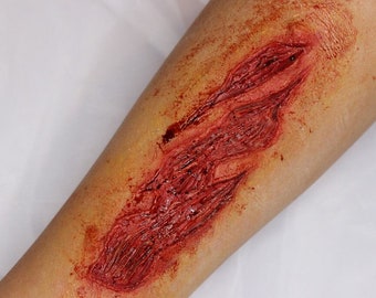 May include: A close-up of a fake, bloody wound on a person's arm. The wound is deep and appears to be a laceration. The wound is covered in fake blood and has a realistic appearance.