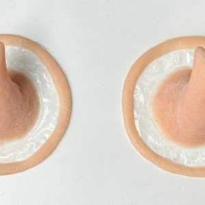 Puede incluir: Dos orejas protésicas de silicona beige con una punta puntiaguda, cada una con un anillo interior blanco.