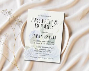 May include: A cream-coloured invitation card for a brunch event, featuring the text "Brunch & Bubbly" and "Emma Smith" in black. The card is decorated with blue and green watercolour floral designs. The invitation is placed on a white plate.