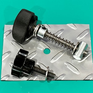 May include: An assortment of hardware components, including black knobs, threaded bolts, a spring-loaded mechanism, and small, round metal discs. The items are arranged on a diamond-plate metal surface against a teal background.