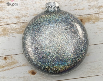 May include: A round, silver glitter Christmas bauble with a metal cap and hanger. The bauble is covered in iridescent glitter, reflecting multiple colours. The words "Craft a Holiday" are in the upper left corner.