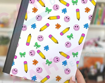 May include: A notebook with a white cover featuring a repeating pattern of yellow pencils, pink disco balls, green bows, orange flowers, and blue flowers. The notebook has a black spine and is held by a hand.