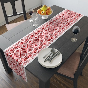 May include: A red and white patterned table runner with a geometric design is displayed on a dark wooden table. The runner features a repeating pattern of snowflakes and other shapes. The table is set with plates, silverware, and wine glasses.