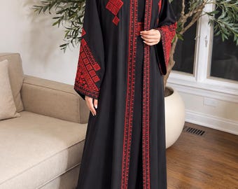 Puede incluir: Una abaya negra larga hasta el suelo con intrincados bordados geométricos rojos en los hombros, mangas y paneles frontales. La prenda tiene mangas largas y anchas y una silueta de corte recto, con el patrón rojo extendiéndose por el dobladillo.