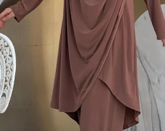 May include: A brown, long-sleeved tunic and matching pants set. The tunic features a draped cowl neckline and an asymmetrical hem. The outfit is made of a soft, flowing fabric. The model is wearing a matching head covering and beige flats.