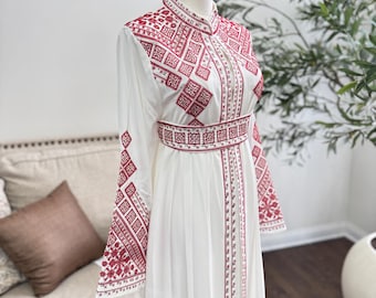 Puede incluir: Vestido blanco con intrincados bordados rojos. El vestido presenta un cuello alto, mangas largas y una falda fluida. El bordado está detallado con patrones geométricos y motivos florales, creando un contraste llamativo con la tela blanca. El vestido se exhibe en un maniquí.