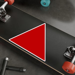 May include: A red triangle sticker with a white border is placed on a black skateboard deck. The skateboard has orange and blue wheels and silver metal trucks. The sticker is a simple geometric shape.