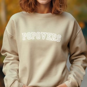 May include: A beige sweatshirt with the word "POPOVERS" printed in white on the front.
