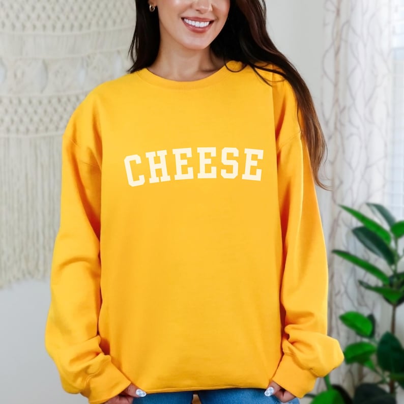 Woman wearing a gold crewneck sweatshirt that says "Cheese" across the front in a collegiate font.