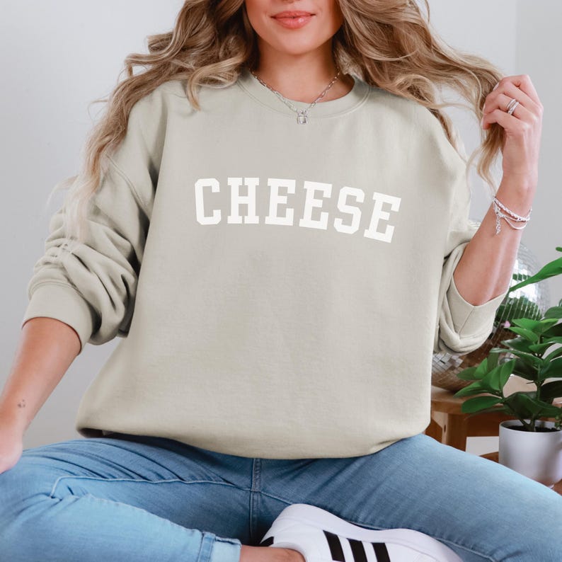 Woman wearing a sand beige colored crewneck sweatshirt that says "Cheese" across the front in a collegiate font.