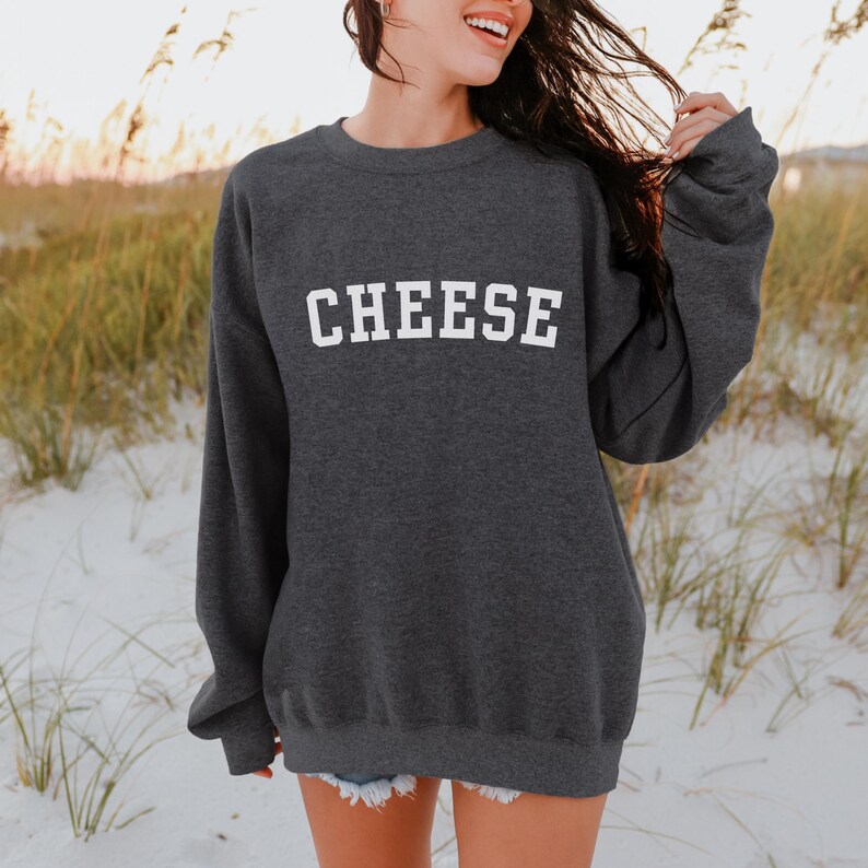 Woman wearing a heather grey crewneck sweatshirt that says "Cheese" across the front in a collegiate font.