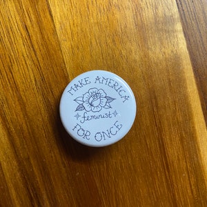 May include: White button with black text that reads "Make America Feminist For Once" and a black outline of a rose.
