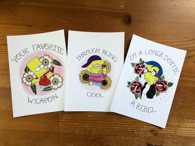 May include: Three illustrated prints featuring characters from the Simpsons cartoon. The first print features Bart Simpson holding a slingshot with the text "Your Favorite Weapon". The second print features Lisa Simpson wearing a purple baseball cap and sunglasses with the text "Through Being Cool". The third print features Milhouse Van Houten wearing glasses and a black leather jacket with the text "I'm a Loner Dottie, A Rebel".