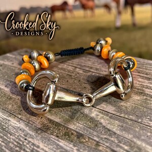 May include: A bracelet featuring a silver-toned metal horse bit centerpiece connected to black cord strands. The cord is adorned with alternating metallic silver and bright orange circular beads, finished with a black adjustable sliding knot closure.