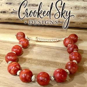 May include: A bracelet featuring large, round, reddish-orange beads interspersed with small, cream-colored, speckled accent beads. The piece includes an adjustable woven cord closure. The text Crooked Sky DESIGNS is visible above the bracelet.