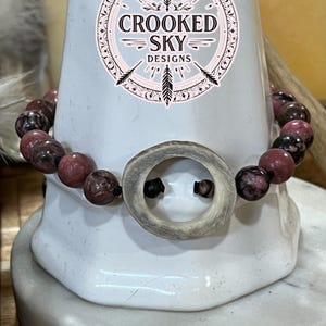 May include: A beaded bracelet featuring pink and brown gemstone beads and a central antler-shaped accent. The bracelet is displayed on a white pedestal, with the "Crooked Sky Designs" logo visible in the background.