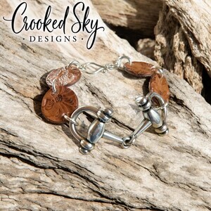 May include: A handcrafted bracelet featuring brown, floral-embossed leather discs and a silver-toned metal clasp. The bracelet includes a decorative bit-style closure. The text "Crooked Sky Designs" is in the upper left corner.