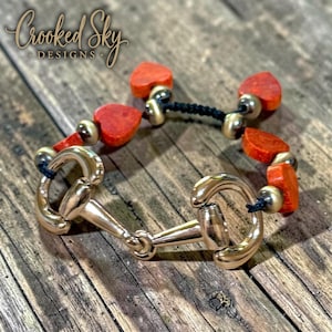 May include: A bracelet featuring a central gold-tone metal horse bit link connected to black cord segments. The design includes small orange heart-shaped beads and metallic gold-tone spherical beads. The text Crooked Sky DESIGNS is visible.