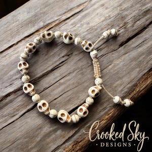 May include: A white skull bead bracelet with a macrame closure. The bracelet is made of white beads with brown skull designs. The bracelet is on a wooden surface with the text "Crooked Sky Designs" in the bottom right corner.