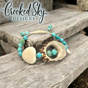 May include: A handmade bracelet featuring turquoise beads, a heart-shaped bone, and a section of antler. The bracelet is strung on a cream-colored cord. The text "Crooked Sky DESIGNS" is visible in the upper left corner.