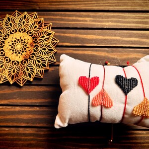 May include: Three friendship bracelets with heart-shaped charms in red, black, and orange. The bracelets are displayed on a white pillow on a wooden surface.