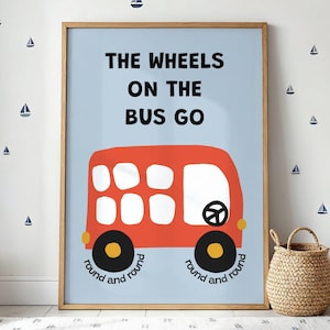 以下が含まれることがあります： 水色の背景に赤いバスのイラストが描かれたフレーム入りプリント。バスの上には「THE WHEELS ON THE BUS GO」の文字、車輪の下には「round and round」と書かれています。壁にはヨットのデカールが飾られています。