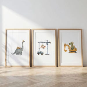 Może przedstawiać: Trzy oprawione wydruki artystyczne z akwarelowymi ilustracjami dinozaurów w sceneriach budowlanych. Jeden wydruk przedstawia dinozaura w kasku, drugi dinozaura obsługującego dźwig, a trzeci dinozaura prowadzącego koparkę.