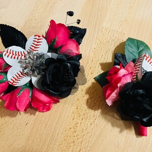 May include: Two baseball themed boutonnieres with red and black roses. One boutonniere features a white baseball with red stitching and a rhinestone embellishment. The other boutonniere features a white baseball with red stitching.