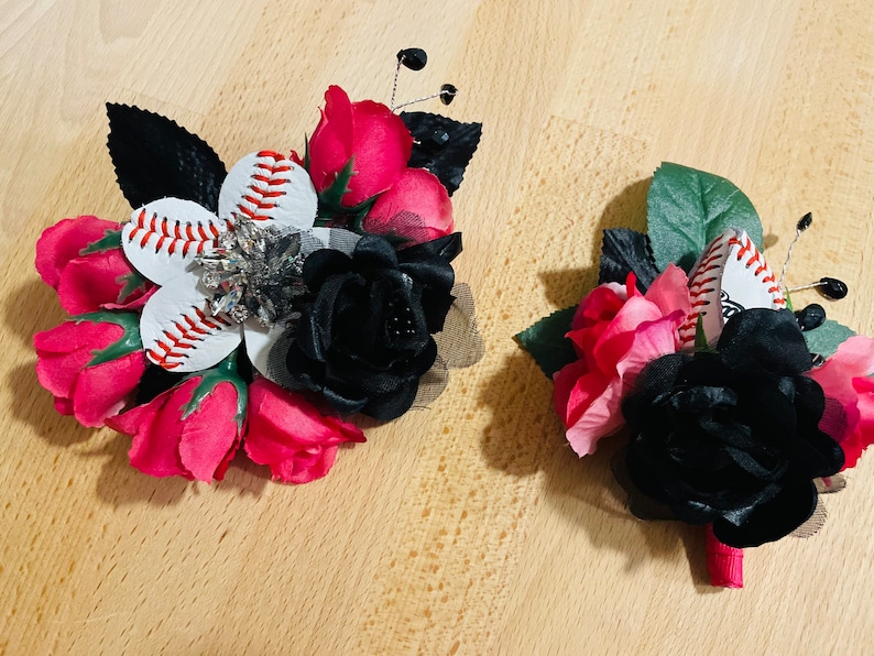 May include: Two baseball themed boutonnieres with red and black roses. One boutonniere features a white baseball with red stitching and a rhinestone embellishment. The other boutonniere features a white baseball with red stitching.