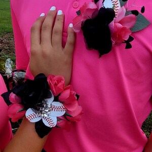 May include: A pink polo shirt with a black and pink flower corsage featuring a baseball. The corsage is pinned to the shirt. A matching wrist corsage with a baseball is also visible.