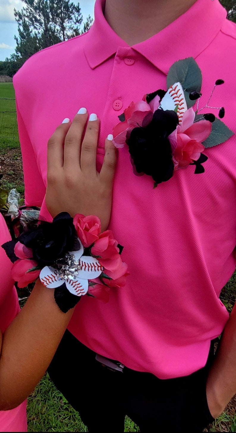 May include: A pink polo shirt with a black and pink flower corsage featuring a baseball. The corsage is pinned to the shirt. A matching wrist corsage with a baseball is also visible.