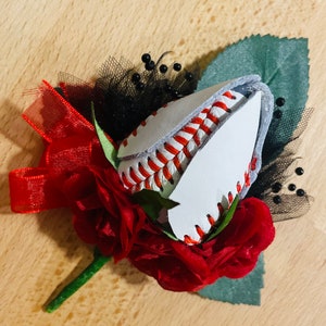 May include: A red rose boutonniere with a baseball stitched into the center. The baseball is white with red stitching and is surrounded by red fabric roses and black netting.