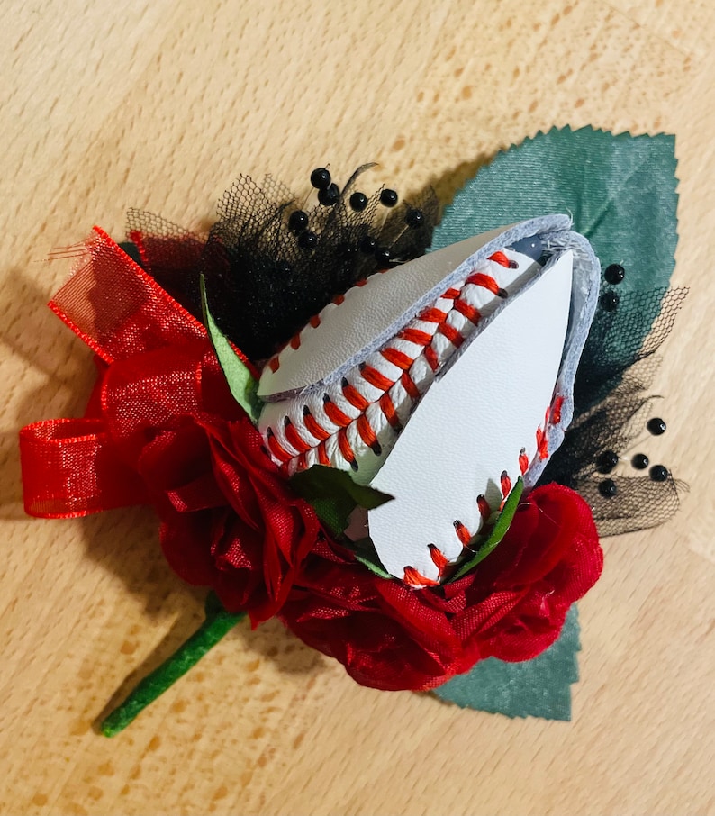 May include: A red rose boutonniere with a baseball stitched into the center. The baseball is white with red stitching and is surrounded by red fabric roses and black netting.