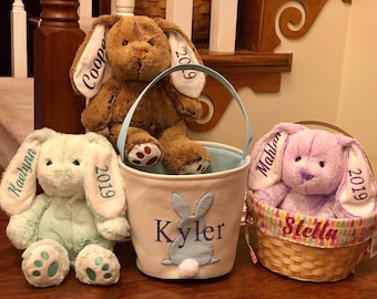 May include: Three plush Easter bunny toys and baskets. A brown bunny sits atop a light blue handled basket with a bunny design and the name "Kyler". A mint green bunny and a purple bunny are also present, each with a name and the year "2019" embroidered on their ears.