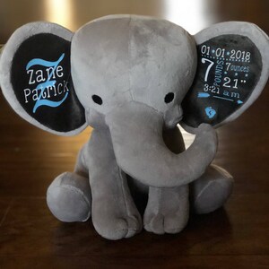 May include: Gray plush elephant stuffed animal with black ears. The ears have embroidered text. One ear says "Zane Patrick" and the other ear says "01*01*2018 7 pounds 7 ounces 3:21 am 21"