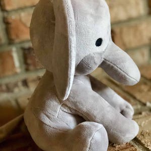 May include: A gray plush elephant toy sitting on a brick surface. The elephant has a large, floppy ear and a black eye.