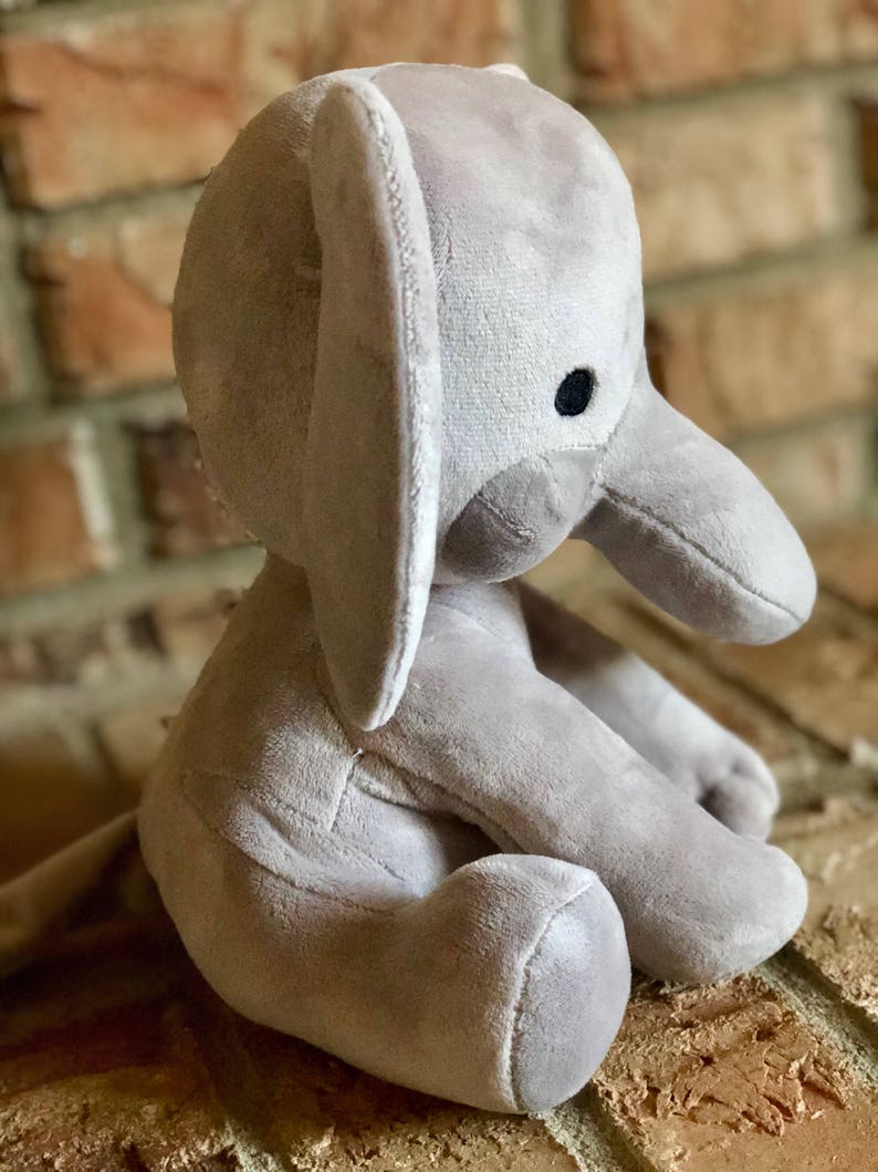 May include: A gray plush elephant toy sitting on a brick surface. The elephant has a large, floppy ear and a black eye.