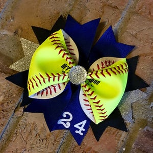May include: A yellow and white softball hair bow with red stitching and a black and blue bow with a silver glitter accent. The bow has a rhinestone in the center and the number 24 on the blue bow.