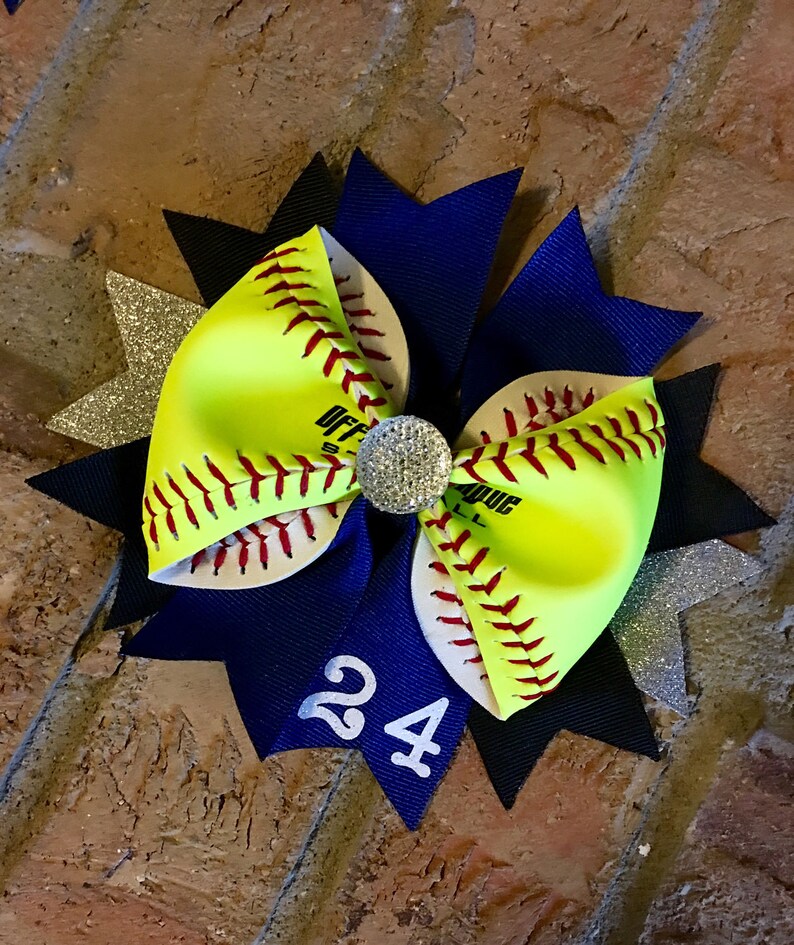 May include: A yellow and white softball hair bow with red stitching and a black and blue bow with a silver glitter accent. The bow has a rhinestone in the center and the number 24 on the blue bow.